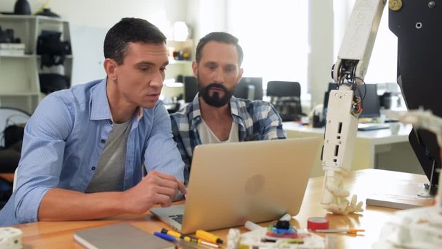 Busy hours. Diligent constructors being concentrated while using a laptop and expressing interest to engineering new robot mockup