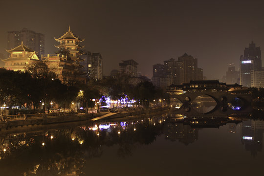 A Skyline Photo of Chendu from the River at Night, With City LIghts Burning Bright and a Bridge reflecting in the still water. Taken in Chengdu, China