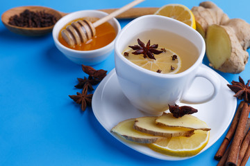 Hot warming ginger tea in white cup with a fine slice of lemon, honey and spices on blue table, close up