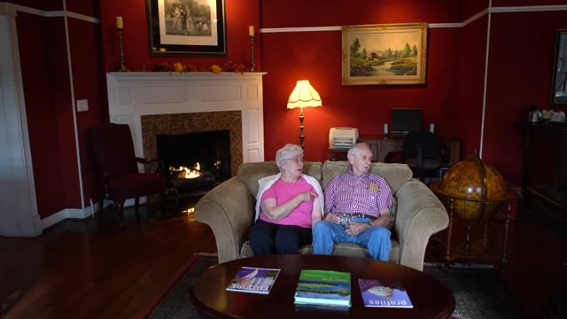 Elderly senior citizen couple sitting on a couch and smiling and talking with a fire in a fireplace in back of them.