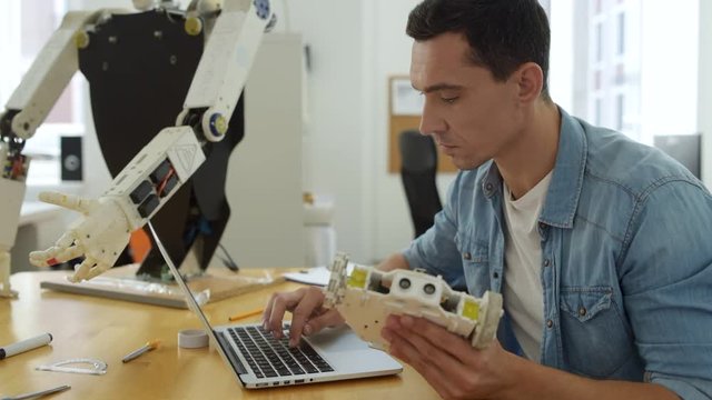 Working atmosphere. Close up of pleasant skilled constructor sitting at the table while using a laptop and holding robot part