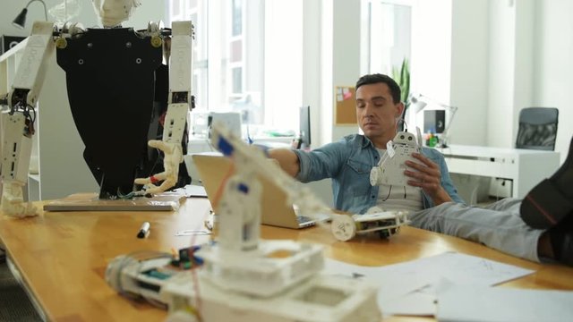Being concentrated. Handsome relaxed constructor sitting at the table while holding robot parts and developing them