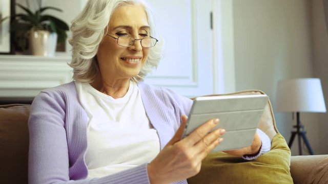 Side portrait view of beatiful old woman in glasses using tablet for surfing in the internet while relaxing on sofa in the living room.