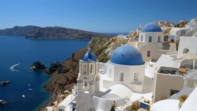 Panning shot of iconic white houses and blue dome churches in Santorini Island, Greece. Sunny day, blue sky