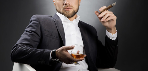 Cognac glass and cigar in hands of man in suit. Dark background