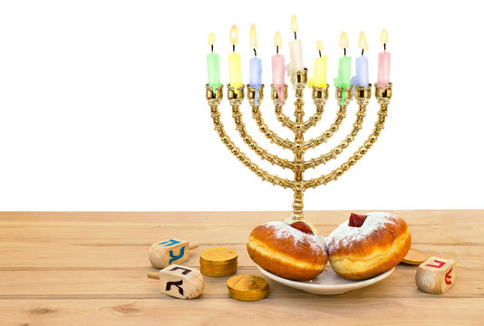 Menorah with candles, donuts, golden chocolate coins and wooden dreidels with red, blue, black letters on wooden table on white background with space for text. Jewish holiday Hanukkah.