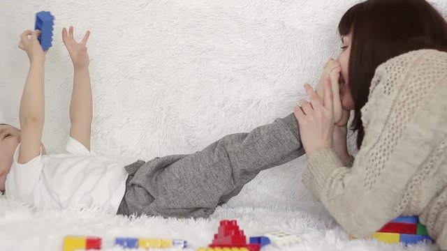 A happy mother kisses her child's legs, lying on a white sofa, against a background of colored toys.