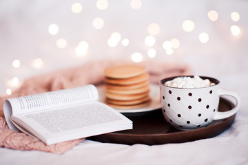 Cup of coffee with marshmallow stay on wooden tray with open book and pancakes in bed over Christmas lights. Good morning. Breakfast.