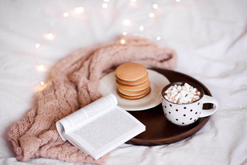 Cup of coffee with marshmallow stay on wooden tray with open book and pancakes in bed over...