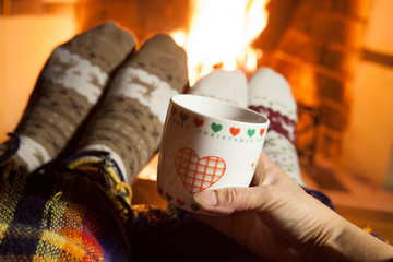 A man and a woman in warm knitted socks with cups of hot drink in front of the fireplace. Red heart on a cup