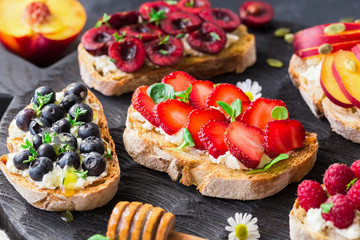 Toasts with goat cheese, honey, strawberries, blueberries, cherries, peaches and raspberries on black wooden cutting board