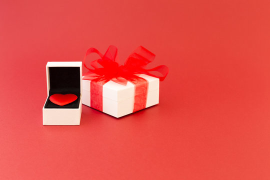 A red velvet heart in a white jewelry box and a white gift box with red ribbon on red background.