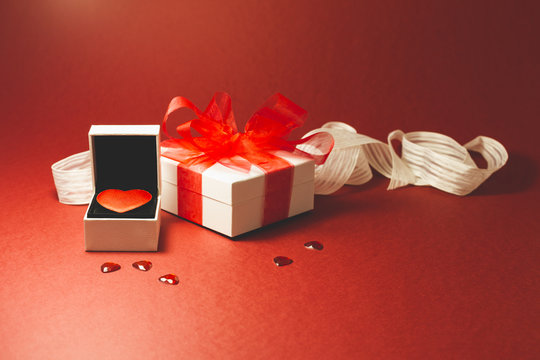 A red velvet heart in a white jewelry box, a white gift box with red ribbon and small shiny red decorative hearts on red background.