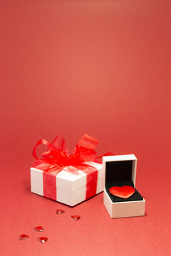 A red velvet heart in a white jewelry box, a white gift box with red ribbon and small shiny red decorative hearts on red background.