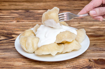 Hand holding boiled varenyky on fork and plate with vareniki on wooden table.