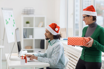 Busy manager in Santa cap working in front of computer monitor while his colleague preparing him xmas surprise