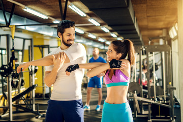 Beautiful attractive couple doing full arm stretch while looking and smiling each other in the modern gym.