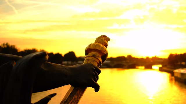 The hand of a sculpture of the famous Pont Alexandre III in Paris. The sun is rising behind the bridge painting the city and the river Seine in orange light.