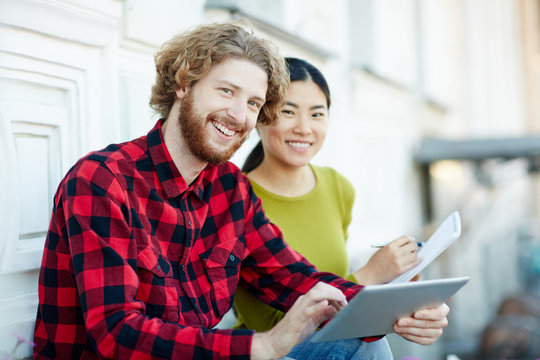 Young man with touchpad and his groupmate carrying out online tasks or test outdoors