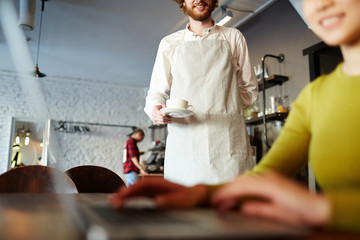 Young waiter in white shirt and linen apron holding cup of tea for his client