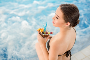 Slim girl with tropical cocktail sitting by swimming-pool with delicate waves of warm water