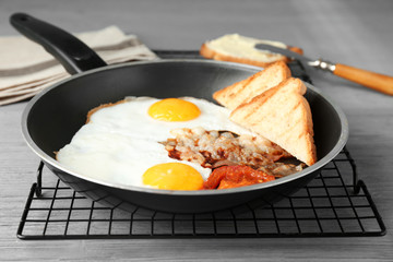 Frying pan with eggs, bacon, toasts and tomato slice on table