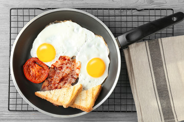 Frying pan with eggs, bacon, toasts and tomato slice on table