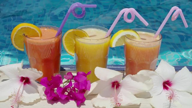Three glasses of vitamin freshly squeezed natural juice with fruit slices and straws on the pool edge at the resort on a hot day. 4K