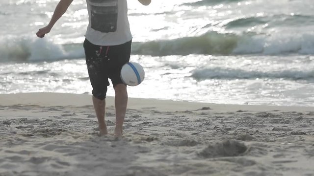 Unrecognizable man kicks football ball at sunset sea beach close up slow motion. Guy fails training alone with soccer ball at sun shine ocean background. Amateur league football player honing skills