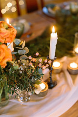 dating, hospitality, comfort concept. tall slim candle that is standing in black holder surrounded by lots of small ones, placed by it on the table with romantic setting of flowers and dishes