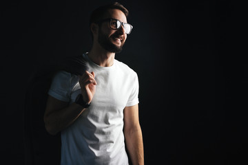 Smiling slender bearded hipster with glasses with a stylish coat on his shoulder wearing white blank tshirt on dark background