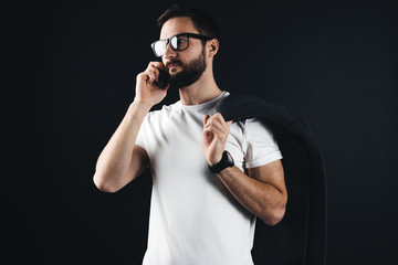 Young bearded handsome man wearing blank t shirt with coat on shoulder. Hipster with glasses posing in white shirt on dark background