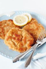 Three golden, pan-fried Wiener Schnitzel made of pork, chicken or veal with traditional breading, two slices of lemon and vintage knife and fork on light blue vintage serving plate in white table with