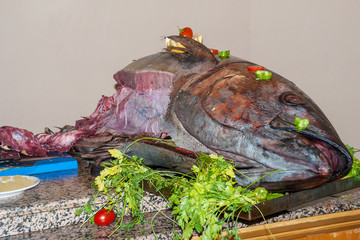 A huge semi-finished tuna lies on a table in a restaurant surrounded by spices and greens