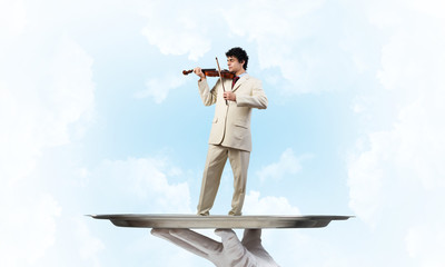 Businessman on metal tray playing violin against blue sky background