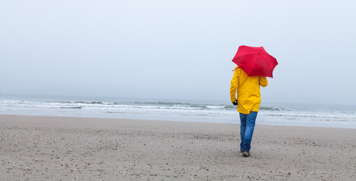 Mann mit gelbem Regenmantel und rotem Schirm am Strand