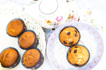 dos platos con magdalenas unas con trozos de chocolate y otras no en platos decorados y una tetera difuminada al fondo
