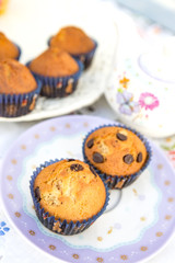 plato con dos magdalenas con trocitos de chocolate en un plato decorado en tonos pastel y al fondo otro plato con magdalenas difuminadas y una tetera al lado.