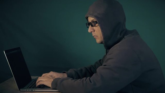 Hoody Man Coding. Profile side view of white man sitting at table and typing at laptop computer. Hacker in glasses and hood crack at dark night. Film look.