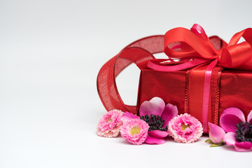 Fototapeta na wymiar Festive gift box with a bow of red color. Lies on a white background. Next to pink flowers.