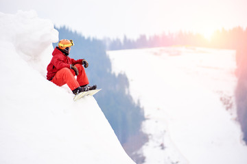 Male snowboarder in a red suit sitting on the top of the snowy hill with snowboard, looking to the...