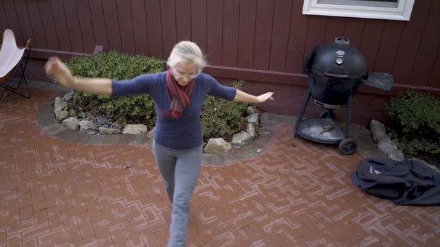 With outstretched arms a mature blonde woman practices middle eastern folk dance on her patio.
