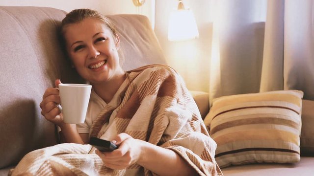 Young woman is sitting in plaide with tea and watching TV at home.

