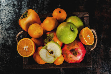 Fresh fruit variety in wooden tray. Oranges, pomegranate, apples, tangerine and persimmon. Dark rusty background, copy space top view