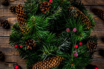 Preparation for xmas holidays. Christmas decorative green wreath with pine cones and red winter berries and christmas tree balls, wooden background, top close view