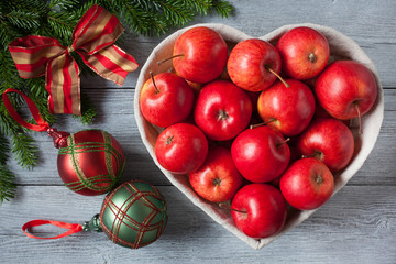 Red apples in a basket in the shape of a heart. Christmas, spruce branches, balls and bow.