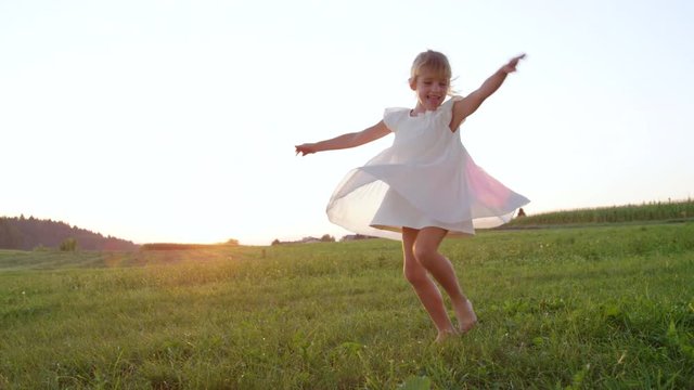 SLOW MOTION CLOSE UP Silly girl dancing and spinning on a green meadow in the late afternoon. Goofy child having fun twirling in a flowing white dress. Young sister dancing barefoot on a grassy field.