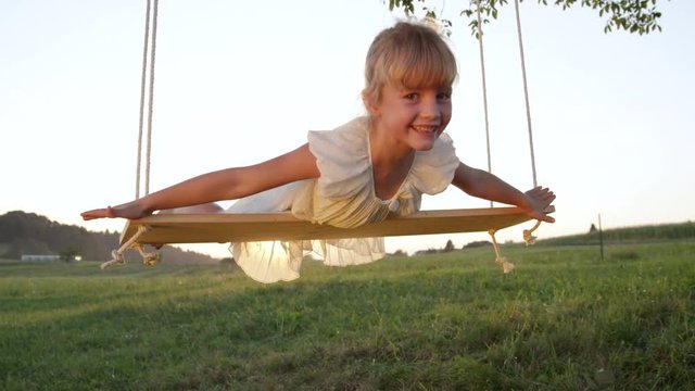 SLOW MOTION PORTRAIT Happy playful child pretending she's flying on a wooden swing. Young silly girl having fun swaying and smiling at the camera. Goofy kid playing pretend and enjoying her summer.