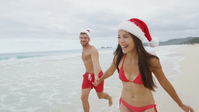 People wearing santa hat - Christmas couple happy having fun on beach travel running on sand in bikini and swimsuit. Asian woman and man holding hands running playing during travel holidays.