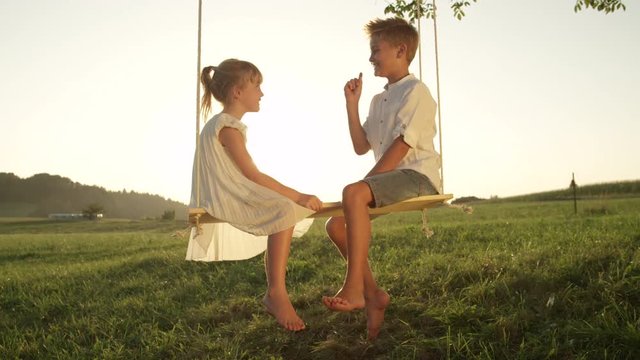 SLOW MOTION CLOSE UP Smiling brother and sister sitting on wooden swing under a tree having fun at sunset. Couple of playful kids facing each other talking on a swing turn attention to the camera